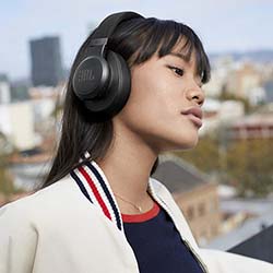 Young woman wearing black over-ear headphones, looking pensive. She stands on a rooftop, cityscape blurred in the background, exuding a calm mood.