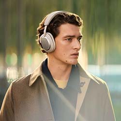 A man wearing large over-ear headphones and a tan coat looks thoughtfully to the side. The blurred background suggests an outdoor urban setting, conveying a serene mood.