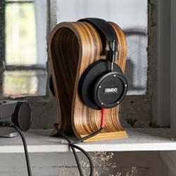 Sleek black headphones rest on a curved wooden stand by a window, casting soft shadows. The scene conveys a modern, minimalist aesthetic.