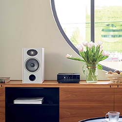 Minimalist room with a wooden shelf, white speaker, small sound system, and vase of tulips by a circular window, creating a serene ambiance.