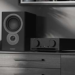 A modern black speaker and audio amplifier sit on a wooden cabinet. Shadows create a sleek, minimalist atmosphere in a home audio setup.