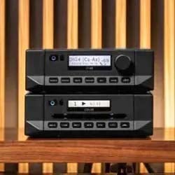 Stacked black audio components with digital displays and button controls, set on a wooden surface. Vertical wood slat background adds a modern touch.