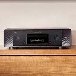 A sleek, black audio receiver with a digital display sits on a wooden shelf. The display shows an audio-related message. The setting is modern and minimalistic.