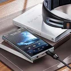 A smartphone with a music player app is on a wooden desk, showing a song playing. Nearby are a pair of headphones and a closed book. The scene suggests a relaxing, musical atmosphere.