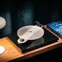 A modern turntable with a sleek white platter and black base sits on a wooden surface. A soft glow suggests a cozy and sophisticated ambiance.