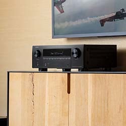 A black audio receiver with a digital display and two knobs sits on a wooden cabinet. Above, part of a wall-mounted TV is visible with an aerial image.