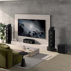 Modern living room setup with a large TV on a wooden panel, surrounded by tall speakers. A green sofa and small table add comfort. Bright, minimalist design.