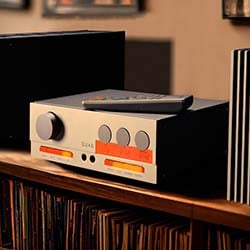 Vintage stereo amplifier on a wooden shelf, surrounded by vinyl records. Warm lighting gives a nostalgic, cozy ambiance, with a remote control on top.
