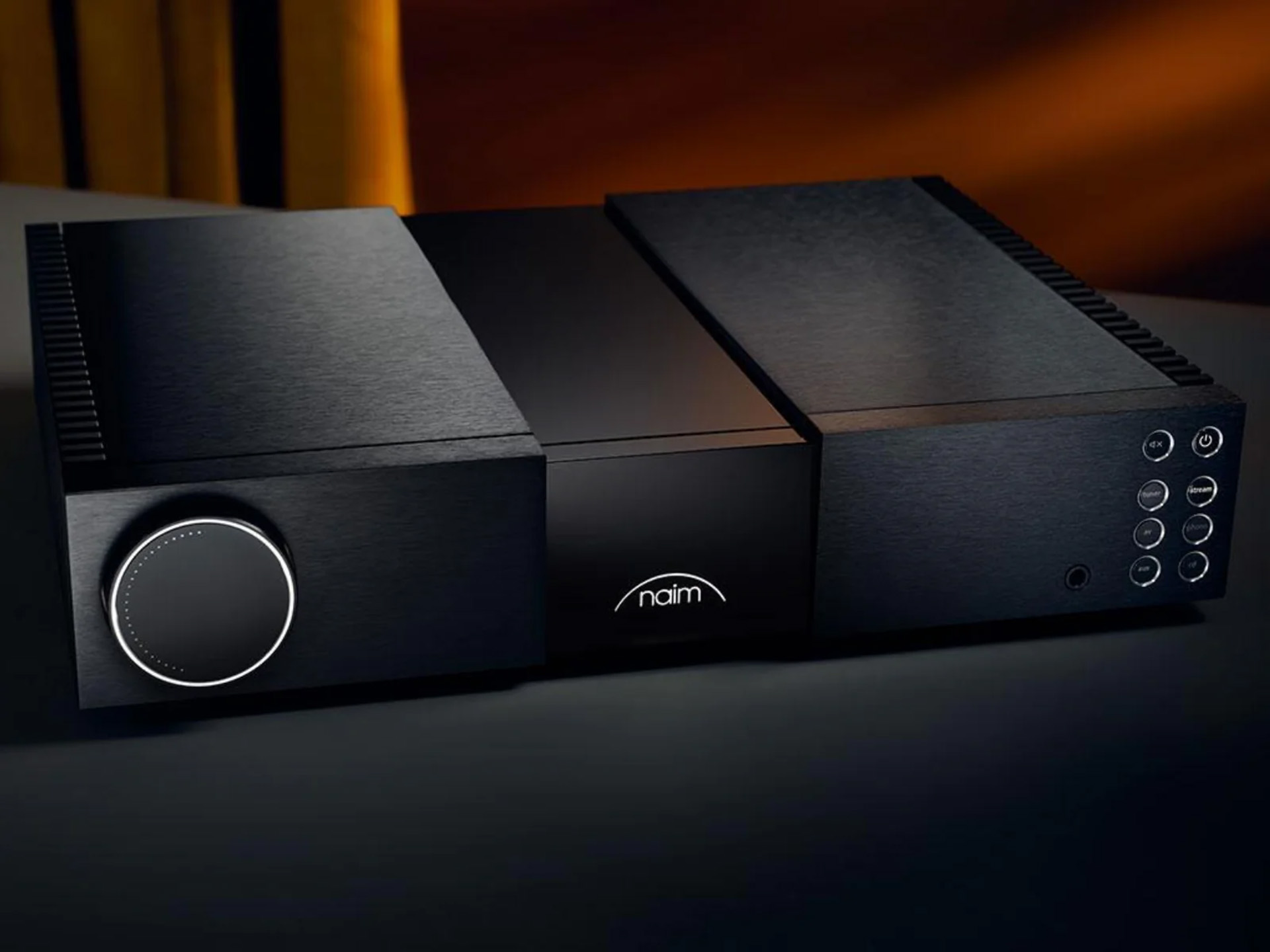 Sleek black audio amplifier with a large circular dial on the left, buttons on the right, and a softly illuminated logo in the center, set against a warm backdrop.