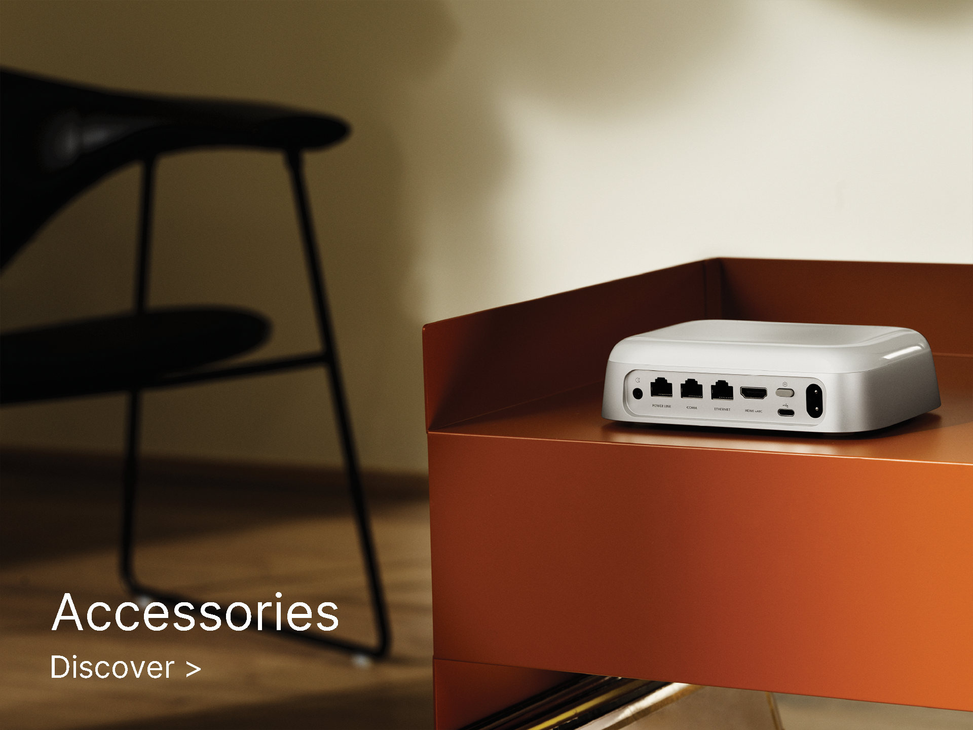 A sleek, silver device with various ports sits on a modern, angular, orange table in a minimalist room. Text reads "Accessories" and "Discover" in the foreground.
