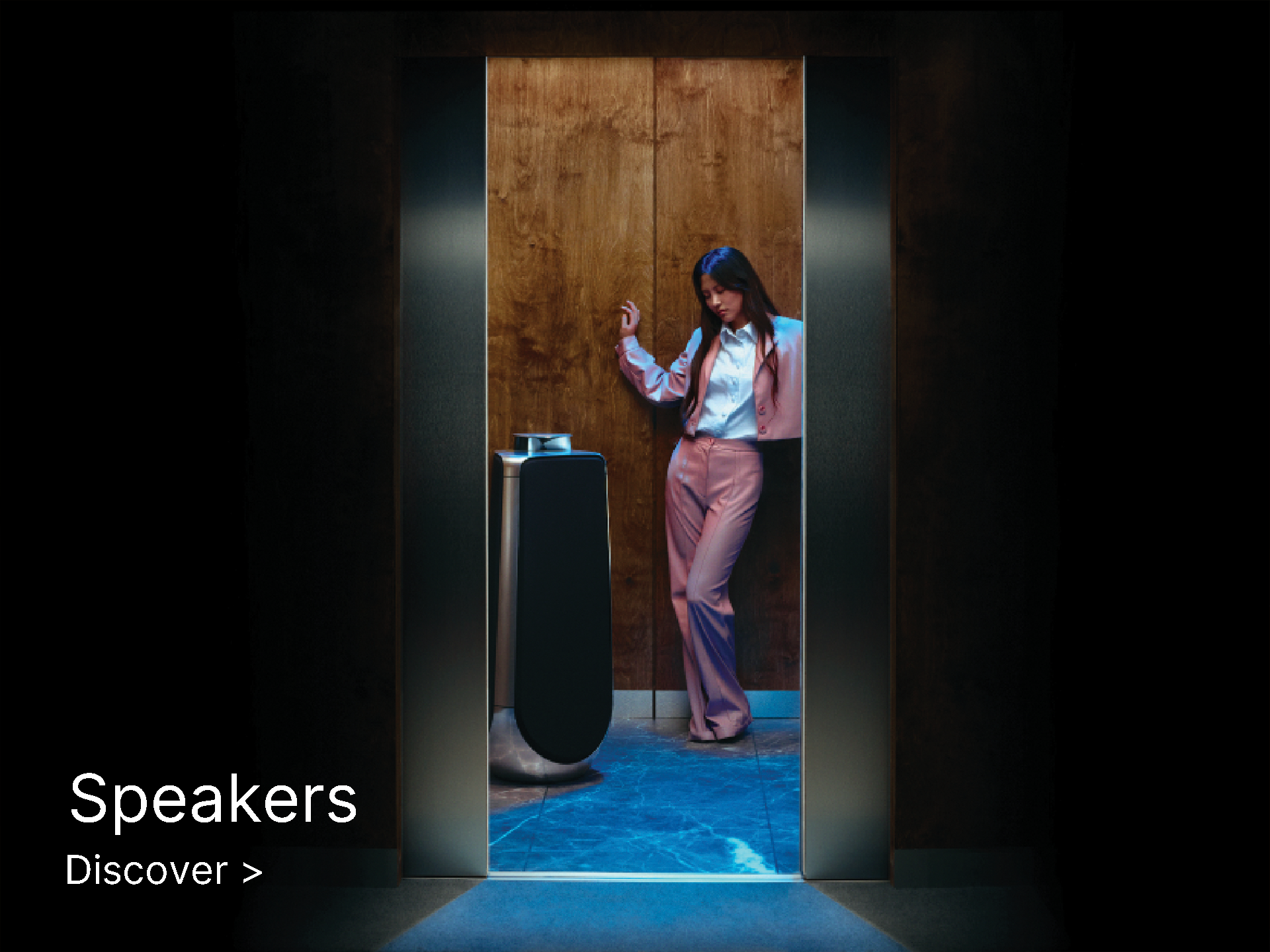 A woman in a pink suit stands beside a modern speaker in a dimly lit room with wooden walls. The scene has a sleek, futuristic vibe. Text reads, "Speakers, Discover."