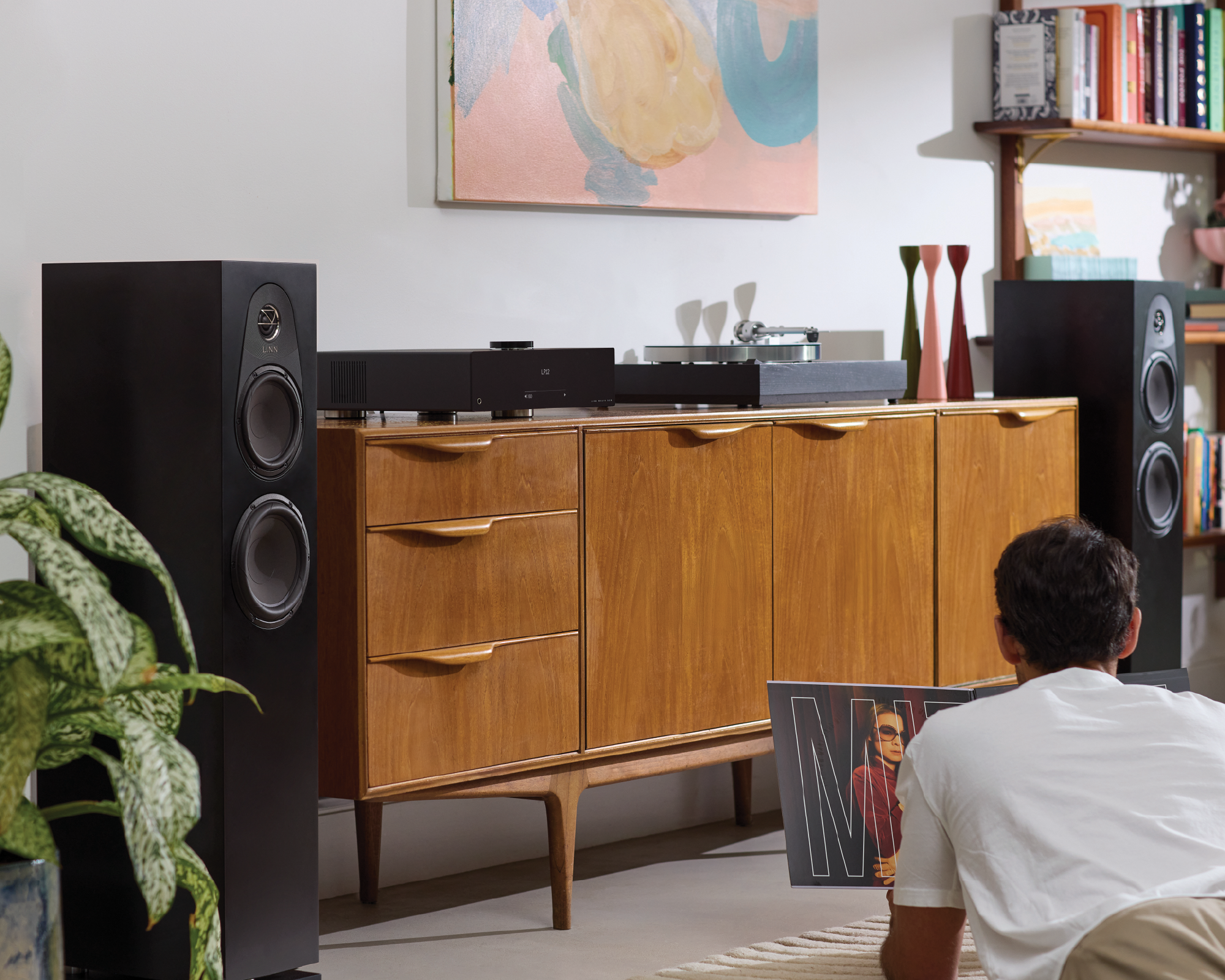 A man sits on the floor holding an album, facing a wooden sideboard with a record player and vinyl records. Large speakers flank the sideboard, creating a cozy, vintage vibe.
