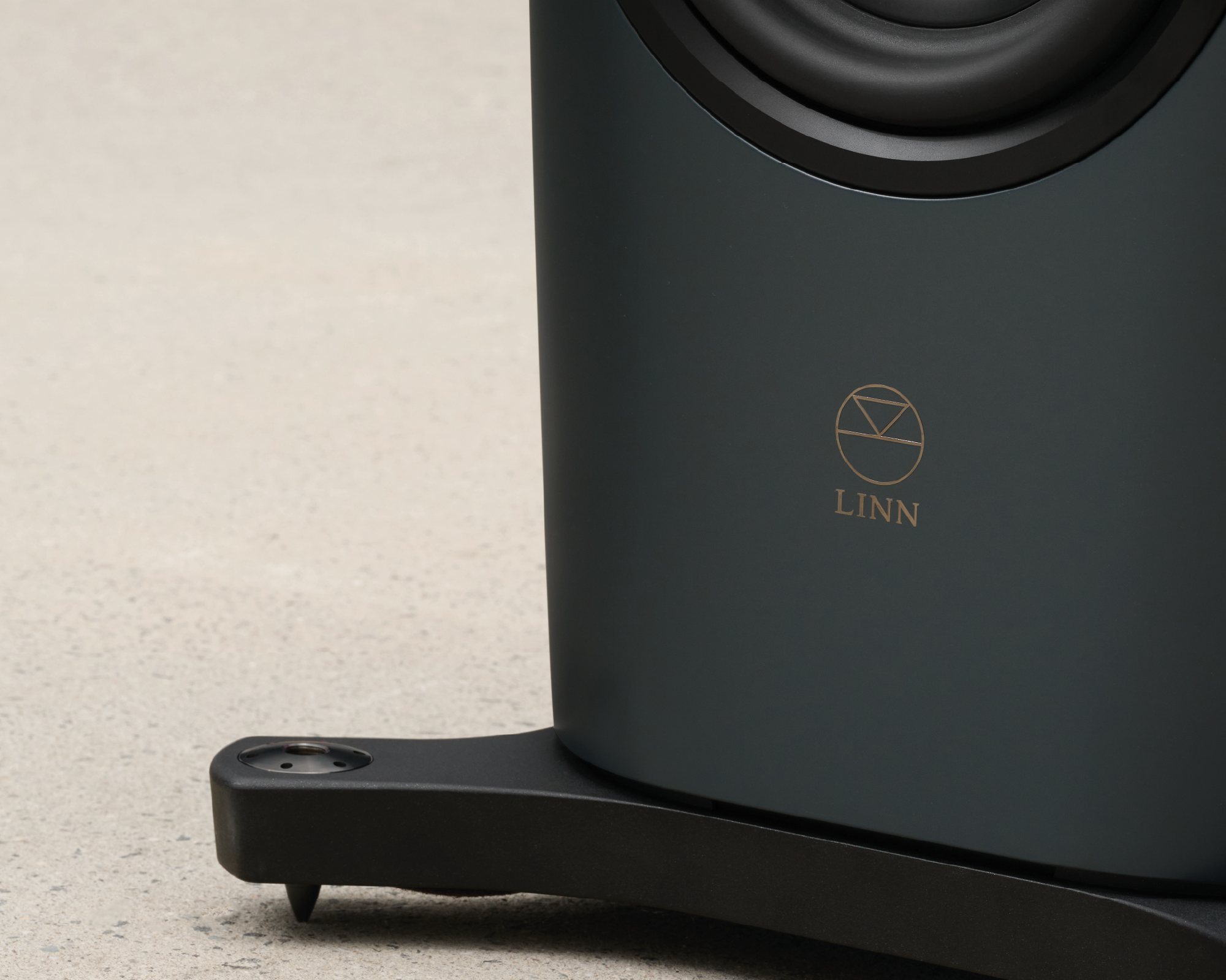 Close-up of a sleek, dark-gray speaker's base, featuring the gold "Linn" logo. It rests on a smooth, light gray surface, conveying elegance.