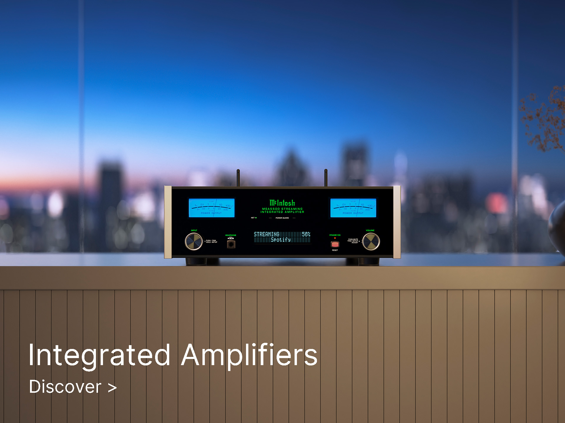 A sleek black audio amplifier with glowing blue meters is displayed on a shelf. Cityscape at dusk in the background. Text: "Integrated Amplifiers, Discover."