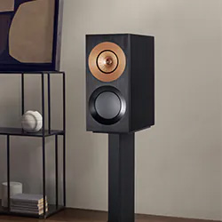 Modern black speaker with a copper-colored tweeter on a stand in a minimalist room. Shelving unit with books and decorative items beside it.