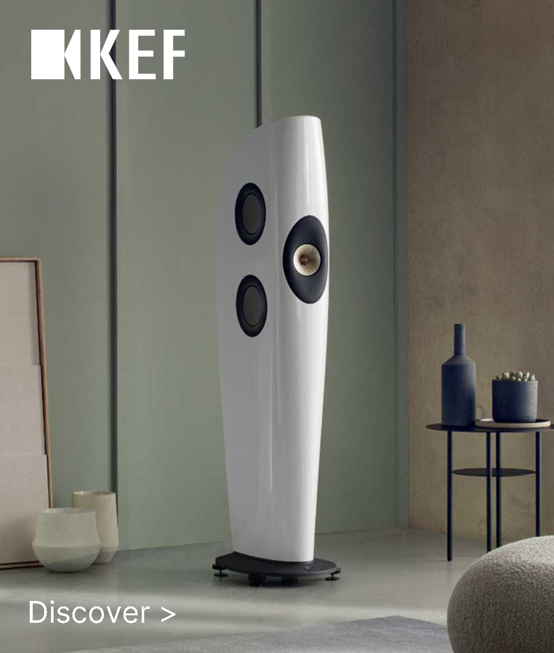 Sleek white KEF speaker with multiple drivers in a modern room. Minimalist décor includes pottery and a small table, conveying elegance and sophistication.