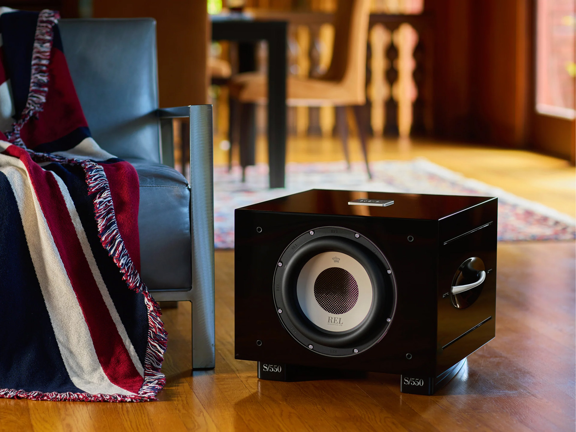 A sleek black subwoofer sits on a wooden floor next to a gray armchair draped with a red, white, and blue striped blanket, creating a cozy atmosphere.