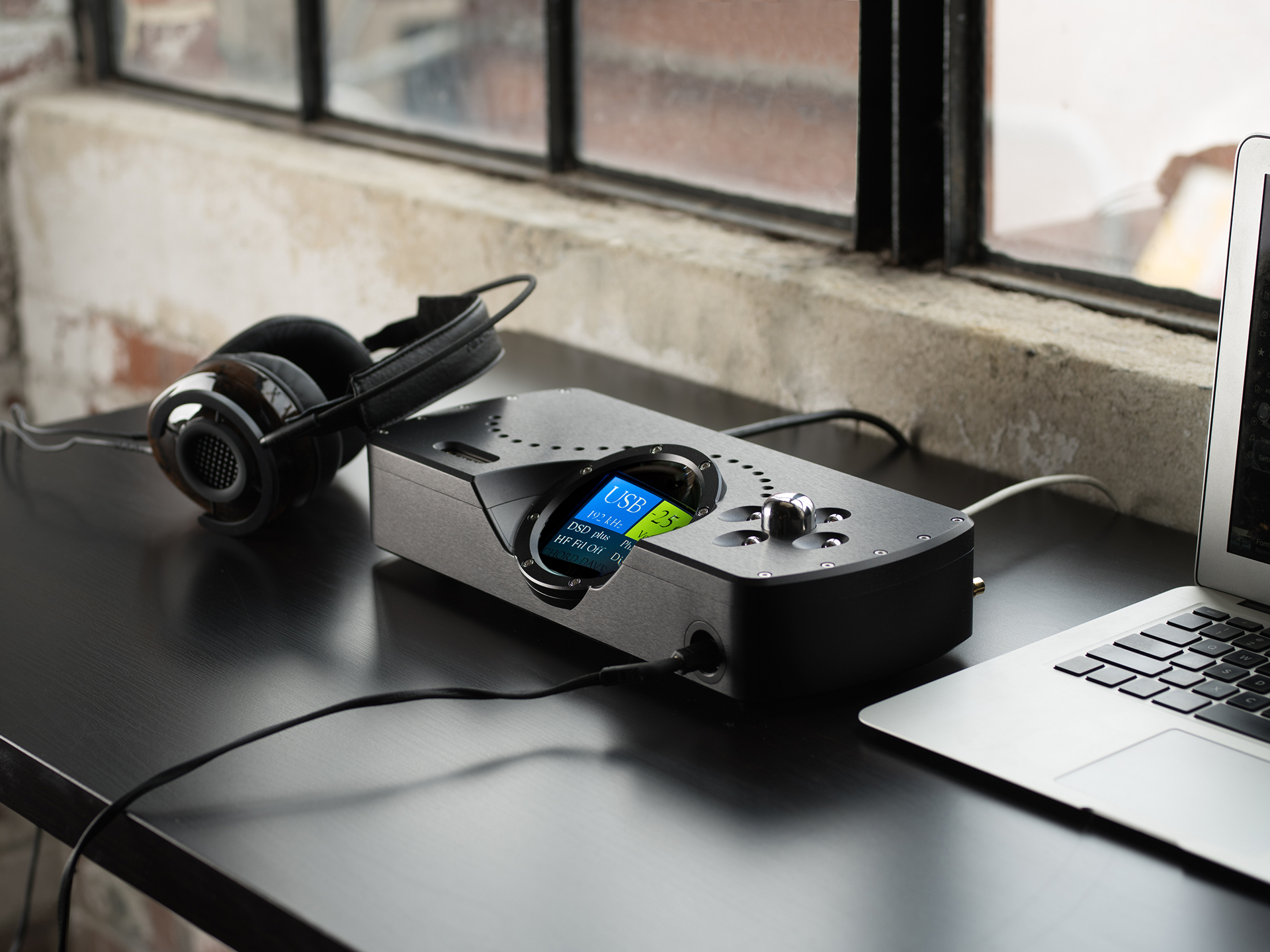 A black headphone amplifier with digital display is on a desk next to headphones and a laptop. Sunlight filters through a window, creating a calm atmosphere.