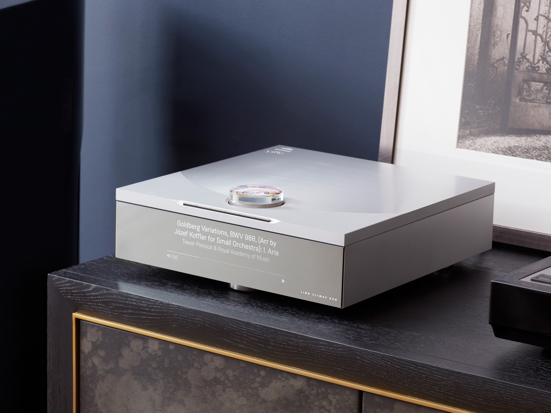 Sleek silver CD player on a dark wood cabinet, displaying "Goldberg Variations" on the front. Modern design with minimal aesthetics, suggesting elegance.