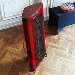 A sleek, red wooden floor speaker with three black circular drivers stands on a herringbone-patterned floor, near a white paneled wall and a blue shelf.