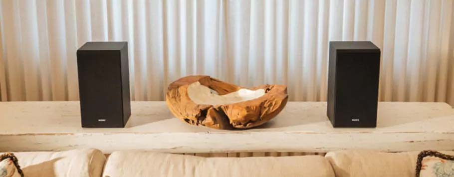 Two black speakers flank a carved wooden bowl on a white shelf, set against light curtains. The setting conveys a modern, minimalist aesthetic.
