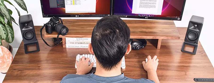 A person sitting at a wooden desk with dual monitors, typing. Speakers, a camera, and books are on the desk. The setting is organized and focused.