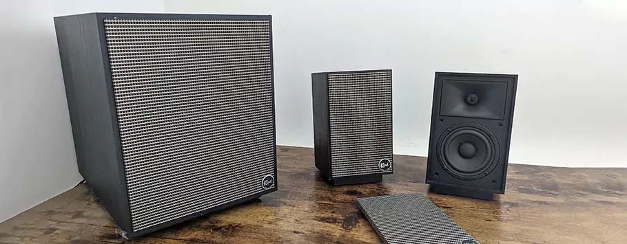 Three speakers with retro designs on a wooden surface. The largest is on the left, two smaller ones beside it, featuring grid-like fronts. One front panel is removed, revealing a speaker cone.