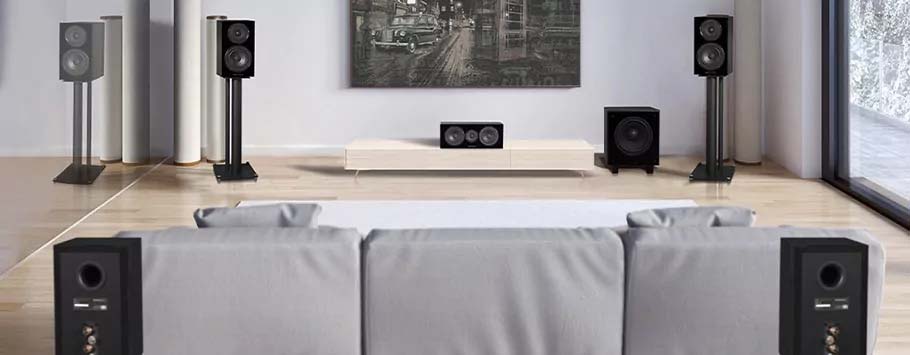 Modern living room with sleek floor-standing speakers, a center speaker, and subwoofer arranged around a large gray sofa, exuding a minimalist vibe.