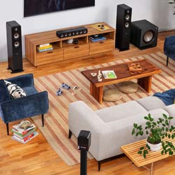 Cozy living room with modern wooden furniture, blue and gray sofas, striped rug, large speakers, and potted plant, conveying a warm, inviting atmosphere.