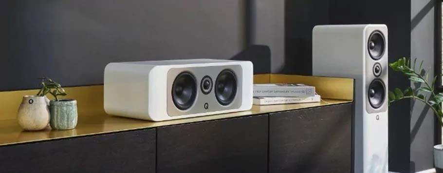 Modern home audio setup featuring a sleek white speaker and a tall speaker with multiple drivers, set on a dark cabinet with potted plants and books.