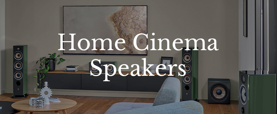 A modern living room with sleek standing speakers flanking a TV mounted on a neutral wall. A gray couch and wooden decor create a cozy, elegant atmosphere. Text reads "Home Cinema Speakers."