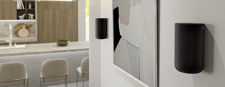 Modern kitchen with minimalist decor. Two black wall-mounted speakers beside abstract art. Neutral tones and wooden cabinetry create a calm ambiance.