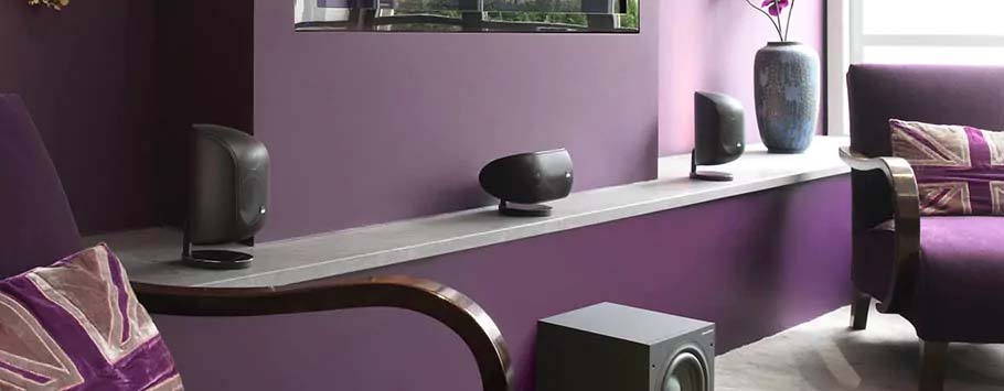 A modern living room with purple walls features sleek black speakers on a ledge, a purple chair with decorative pillows, and a large vase with flowers.