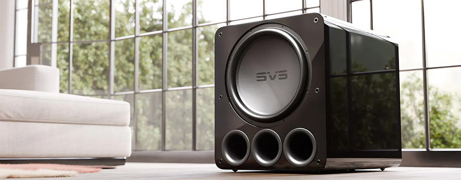 A black SVS subwoofer with three front ports is placed on a wooden floor in a bright room. Large windows and a light-colored sofa are in the background.