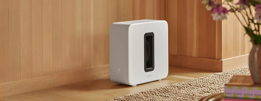 A modern white subwoofer sits against a wooden wall on a carpeted floor. A vase with flowers and a stack of books are partially visible in the foreground.