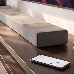 A sleek, modern soundbar on a wooden shelf next to a smartphone. Sunlight creates a warm ambiance, highlighting the minimalist design.