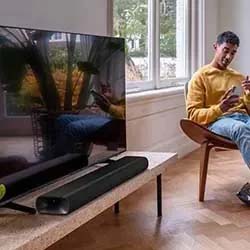 A man in a yellow sweater sits relaxed in a modern chair, looking at his phone. A TV with a soundbar is on a stand nearby, in a bright, airy room.