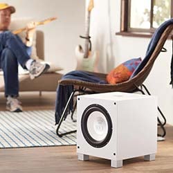 A white subwoofer sits on a wooden floor in a cozy room. A person plays guitar on a beige couch, adding a relaxed, musical vibe to the space.