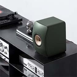 A modern dark green speaker with a copper center is on a sleek black shelf. Nearby, a turntable and audio equipment add a stylish, high-tech vibe.