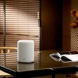 Smart speaker on a sleek table in a cozy room with soft lighting. A chair and open book nearby; blinds partially cover the window, creating a warm ambiance.