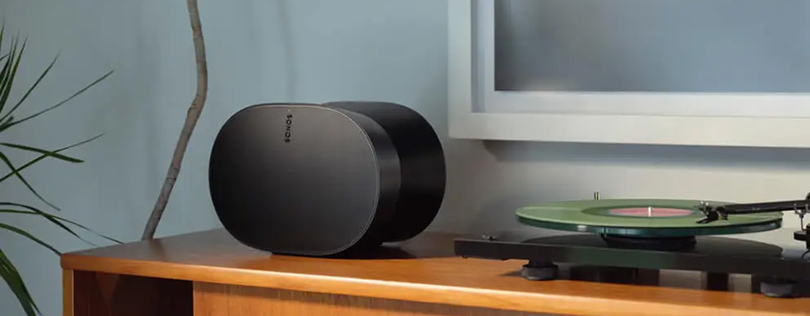 A sleek black speaker labeled "Sonos" sits on a wooden shelf next to a green vinyl record on a turntable. A modern, minimalist setting with a relaxed vibe.