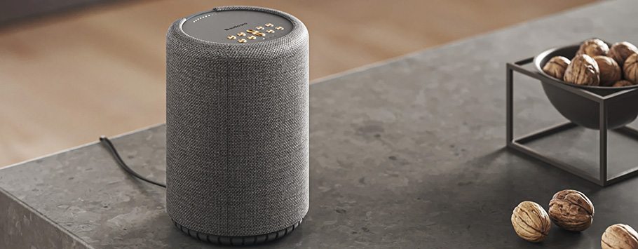 A gray cylindrical smart speaker sits on a marble countertop, near a metal bowl filled with walnuts. The scene conveys a modern and cozy atmosphere.