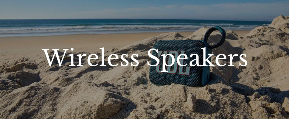 Portable wireless speaker on sandy beach with ocean waves in the background; text "Wireless Speakers" overlays the image, conveying a relaxed vibe.