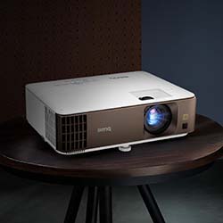 A sleek white and brown projector is placed on a round wooden table. The lens is lit, casting a subtle blue light, set against a minimalist dark background.