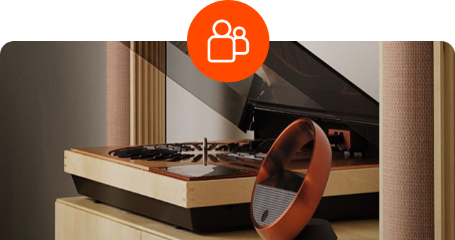 A high-end wooden turntable with an open dust cover sitting next to a circular metallic controller on a light wood console, with a circular orange people icon overlaid at the top center.
