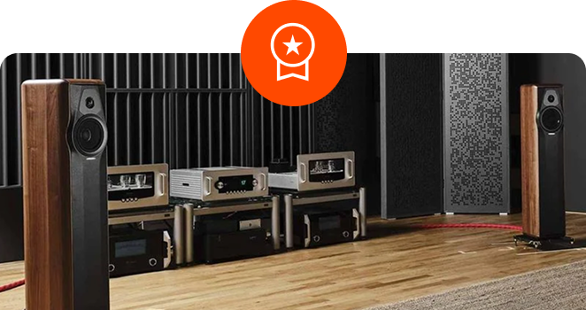 Two tall wooden floor-standing speakers flanking high-end audio amplifiers on a low rack in a listening room with wooden floors, with a circular orange star ribbon icon overlaid at the top center.