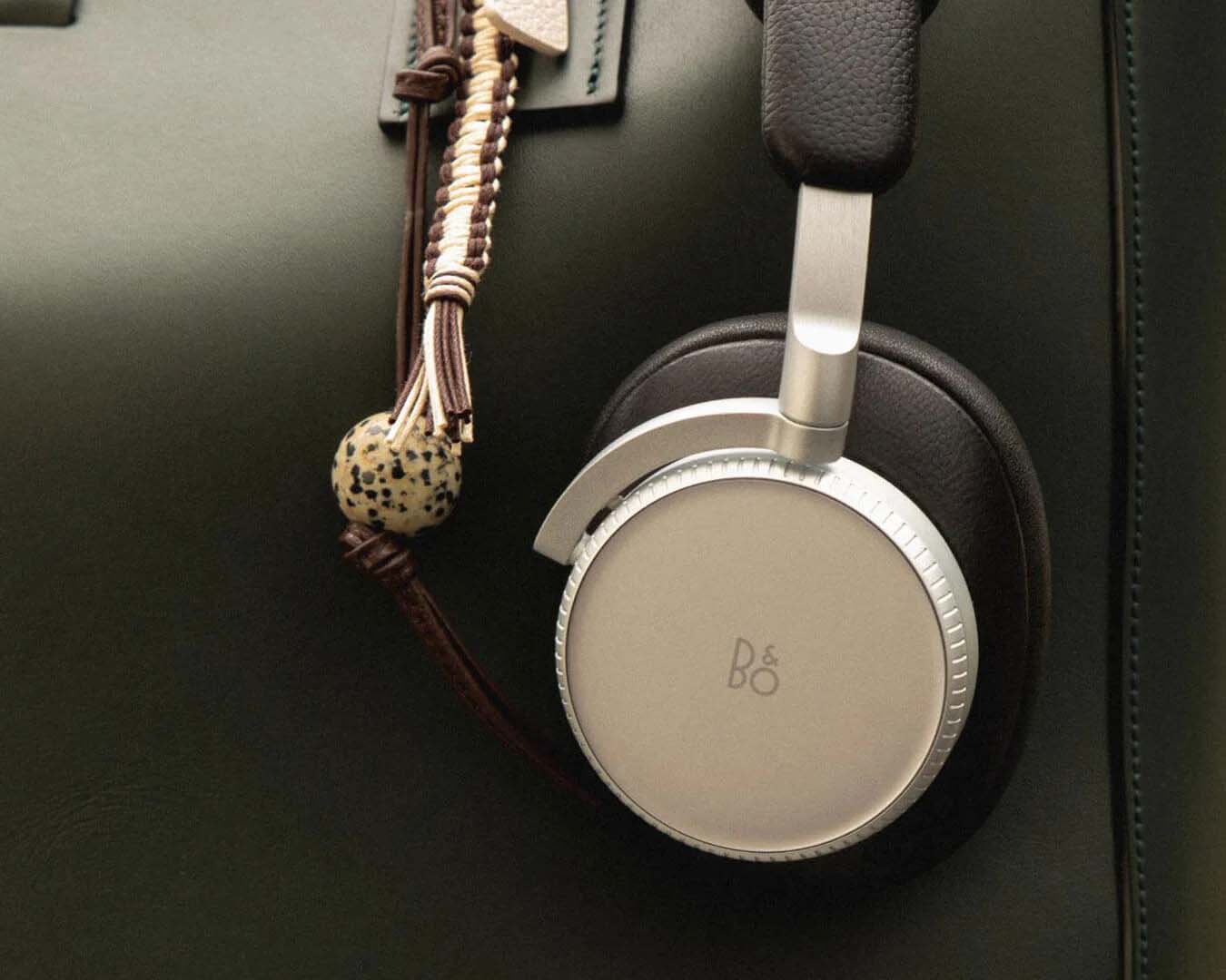 A close-up shot of a silver and dark brown Bang & Olufsen over-ear headphone ear cup resting against a dark leather surface, alongside a braided cord accessory featuring a speckled dalmatian jasper bead.