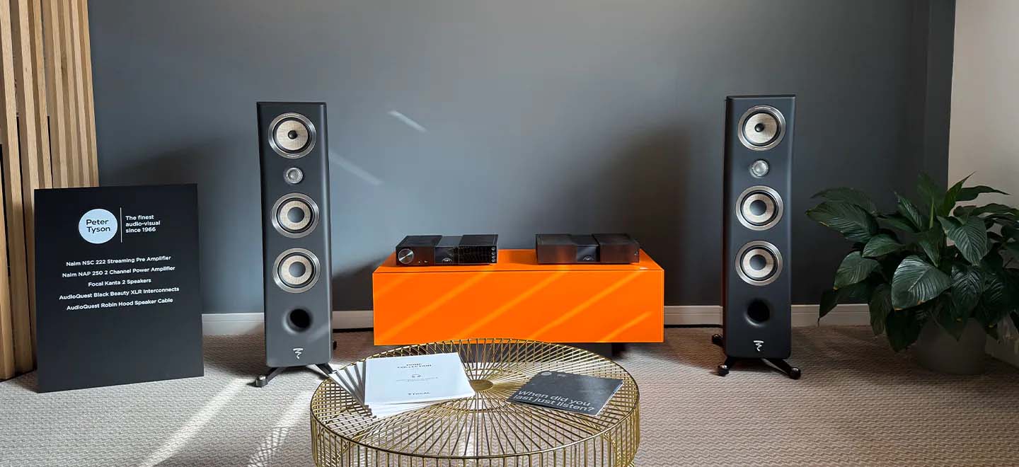 A modern audio setup features two tall black speakers on either side of a bright orange console. A sleek black sign on the left describes product features. A stylish, round, gold table in the foreground holds a brochure. A lush green plant on the right adds a touch of nature. The room has a calm, sophisticated ambiance.