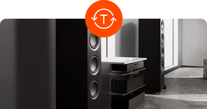 A pair of tall, black floor-standing audio speakers positioned in a modern room, with a round orange badge containing a "T" and circular arrows overlaid at the top center.
