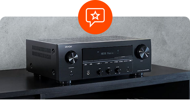 A black Denon stereo network receiver displaying "HEOS Music" on its front panel screen, sitting on a dark wooden surface against a grey wall, with a circular orange chat bubble star icon overlaid at the top center.